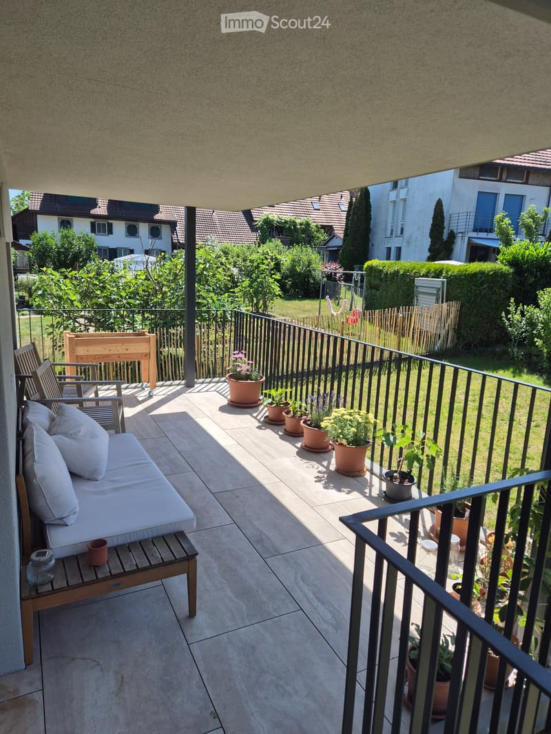 Balcony with a sofa, chairs, a small table, and several potted plants, overlooking a garden area with trees and houses.