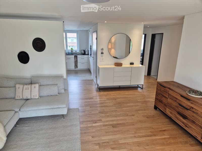 Modern living room with grey sofa, wooden floor, white cabinet, round mirror, and adjacent kitchen area with white cabinets and appliances.