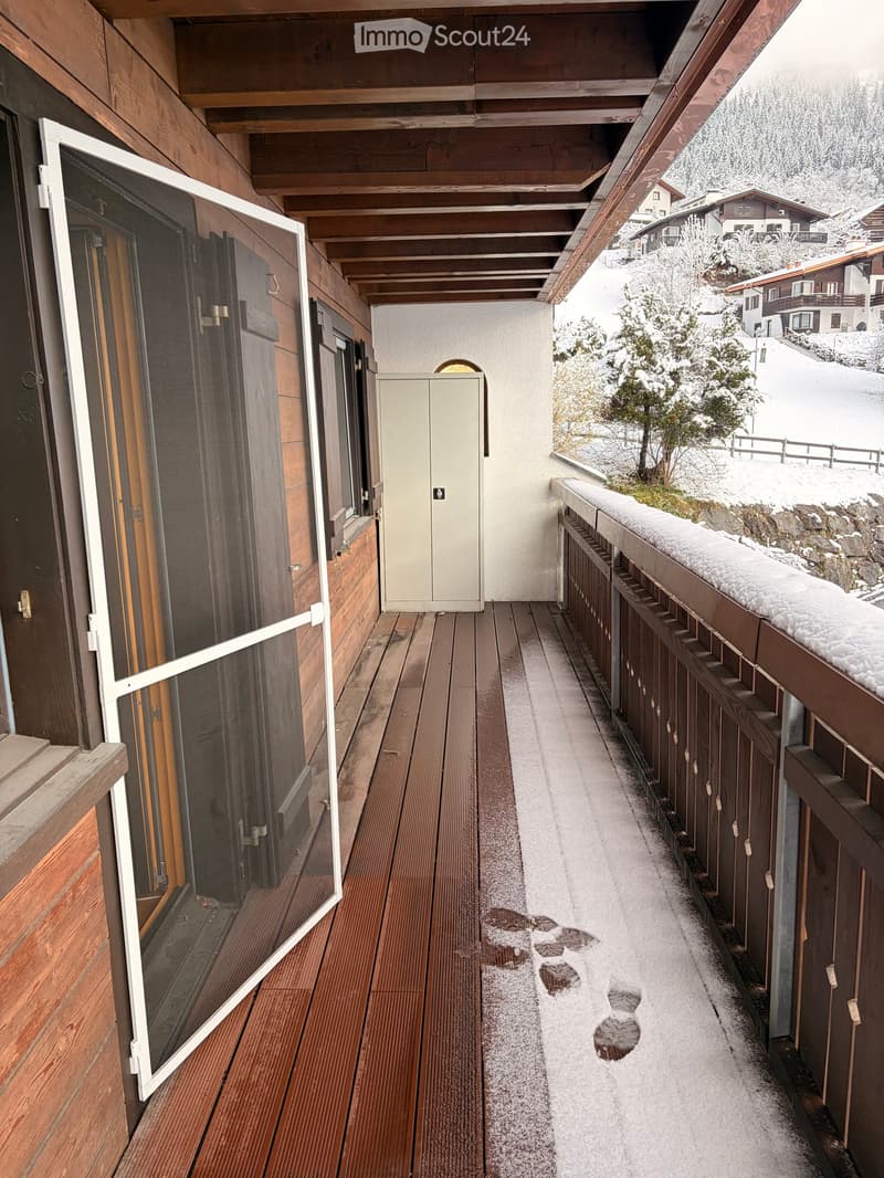 Wooden balcony with snow-covered railing and pathway, overlooking a snowy landscape with houses and trees.