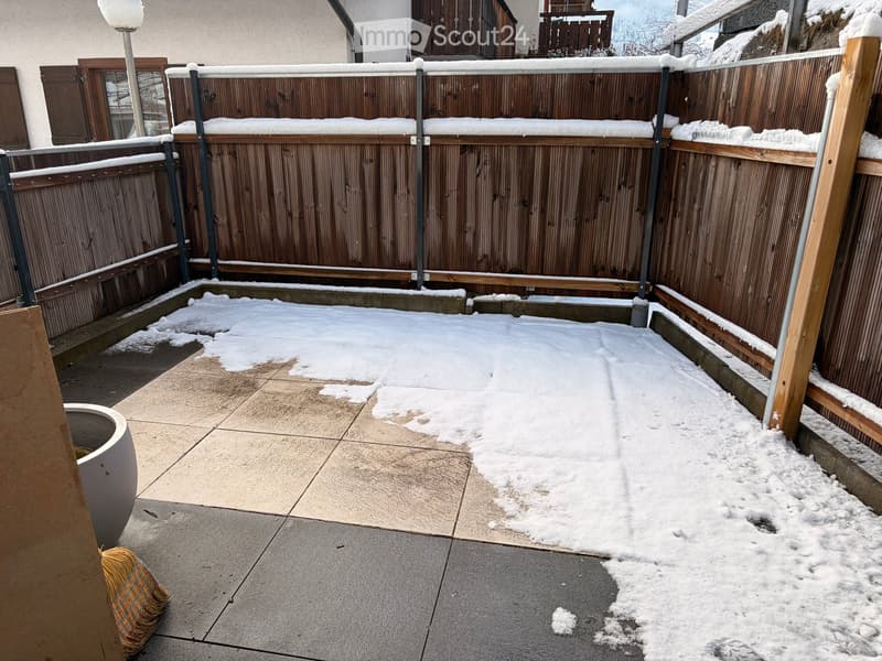 A small outdoor patio area with a wooden fence, snow patches, and a broom leaning against the wall.