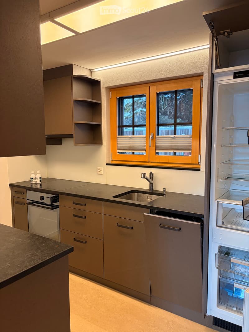 Modern kitchen with grey cabinets, stainless steel appliances, and a window with blinds. Features a sink, faucet, refrigerator, oven, dishwasher, and two bottles on the counter.