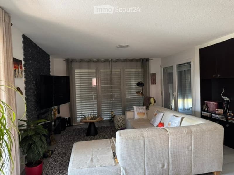 Modern living room with a light-colored sofa, ottoman, coffee table, television, potted plant, and wall-mounted lamp. The space features large windows with blinds and a sleek, contemporary design.