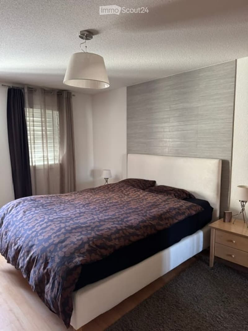 Modern bedroom with a large bed, nightstand, lamp, and curtains. The room features a sleek headboard and a contemporary design.