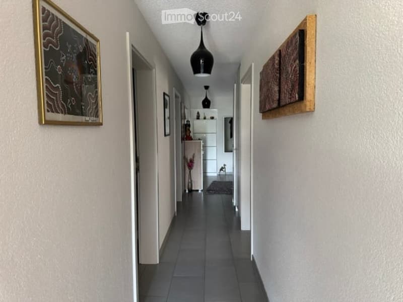 A narrow hallway with tiled flooring, white walls, and modern black pendant lighting. Artwork is displayed on the walls, and a small table with a vase of flowers is visible at the end of the corridor.