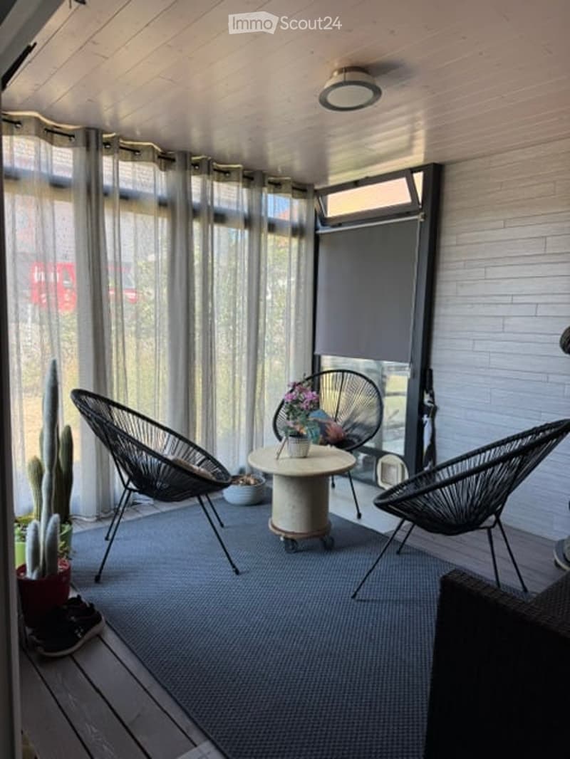 A modern, enclosed balcony with large windows, sheer curtains, and a sleek design. Features include a round table with a vase of flowers, two black wicker chairs, and a small cactus plant in a pot.