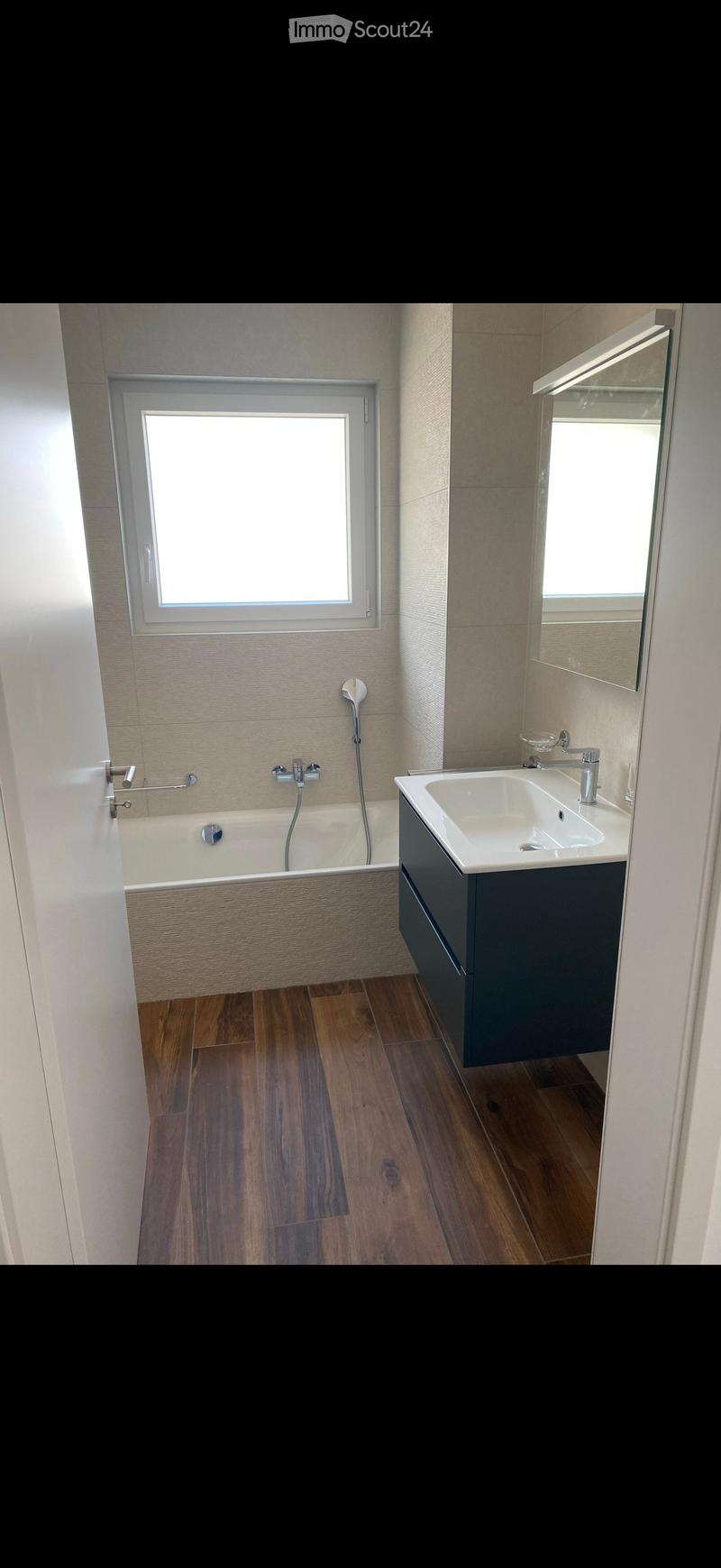 Modern bathroom with a bathtub, sink, mirror, and dark cabinet. Features wooden flooring and a window above the bathtub.