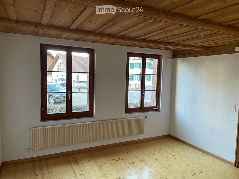 Empty room with wooden floor, white walls, wooden ceiling, two windows, and a radiator below one window.