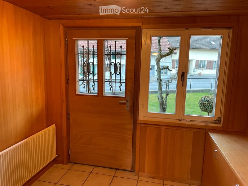 Wooden hallway with a door leading outside, tiled floor, radiator, and window showing a view of the garden and neighboring house.