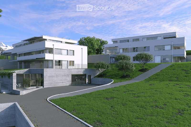 Modern multi-story residential building with clean lines, large windows, and a garage entrance. The structure is set on a raised ground floor with a landscaped green area and pathway leading up to the building.