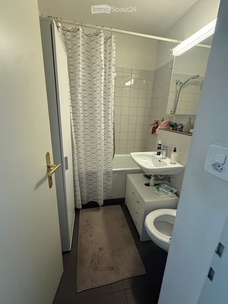 Modern bathroom with white tiles, bathtub, sink, toilet, mirror, and shower curtain. Includes a small cabinet and a bathroom rug on the floor.