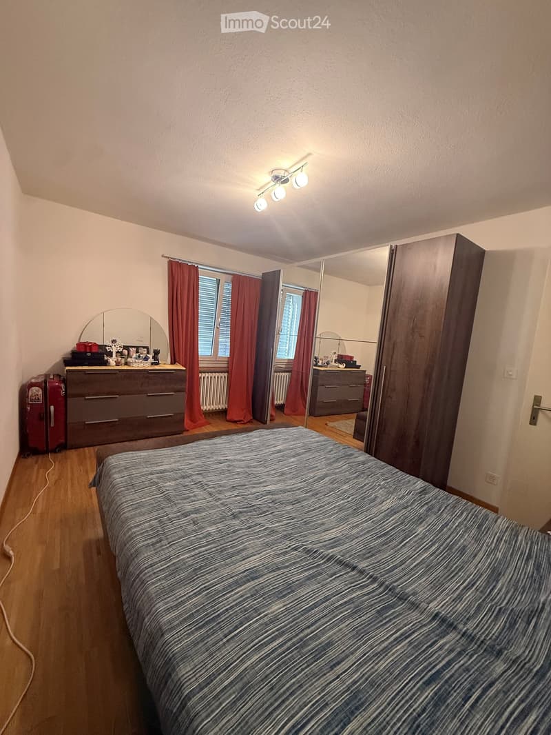 Cozy bedroom with wooden flooring, a large bed with striped bedding, a wardrobe with mirrored doors, a dresser, and a suitcase near the window.