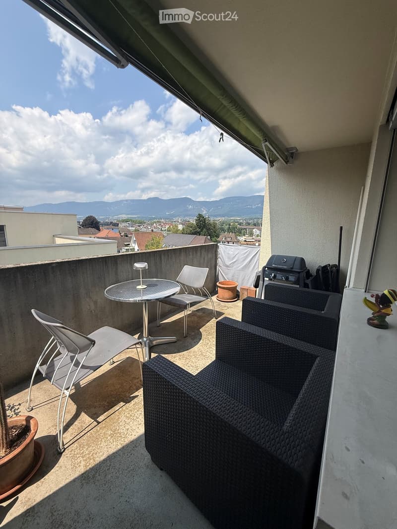 Balcony with a view of mountains, equipped with a round table, chairs, a sofa, and a grill.