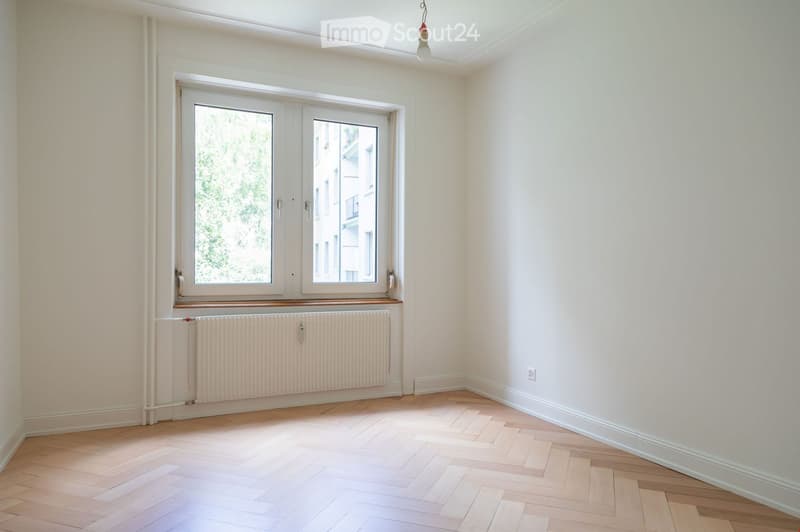 Empty room with light-colored wooden flooring, white walls, and a large window with white frames. A radiator is positioned below the window, and a ceiling light fixture is visible.