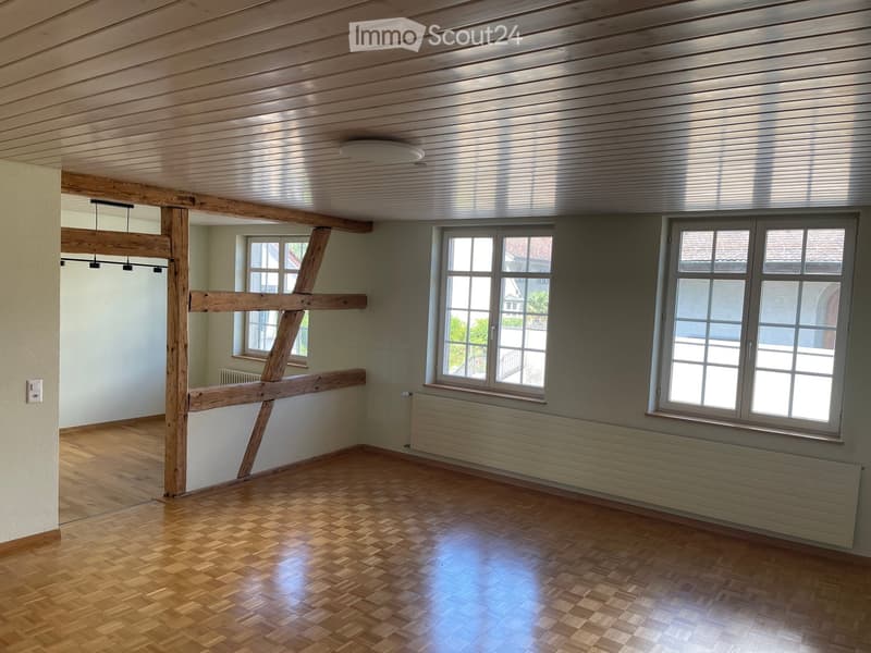 Empty room with wooden floor, exposed wooden beams, and large windows letting in natural light.