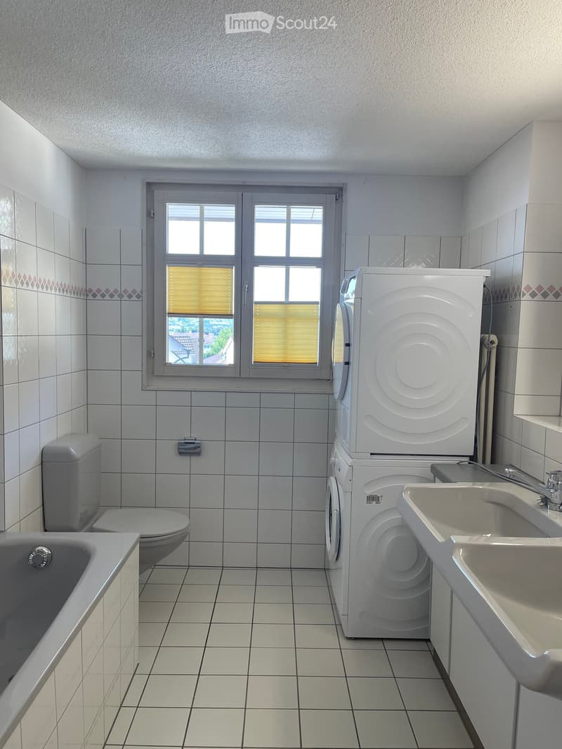 Bathroom with tiled walls and floor, featuring a bathtub, toilet, sink, and a stacked washing machine and dryer unit.