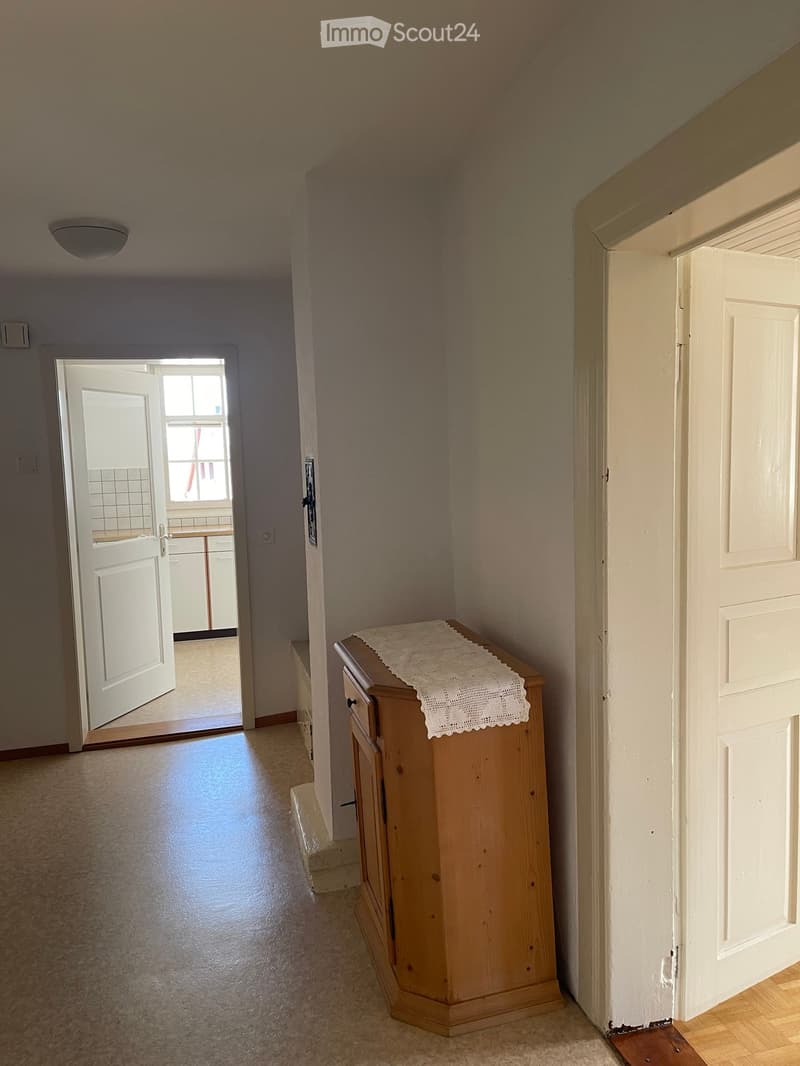 A narrow hallway with light-colored walls, carpeted floor, and a small wooden cabinet with a white cloth on top. An open door leads to a room with a window and a tiled wall visible.