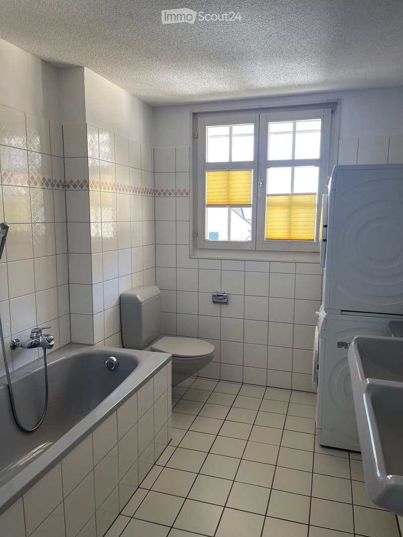 Bathroom with tiled walls and floor, featuring a bathtub, toilet, washing machine, and sink. A window with blinds allows natural light to enter.