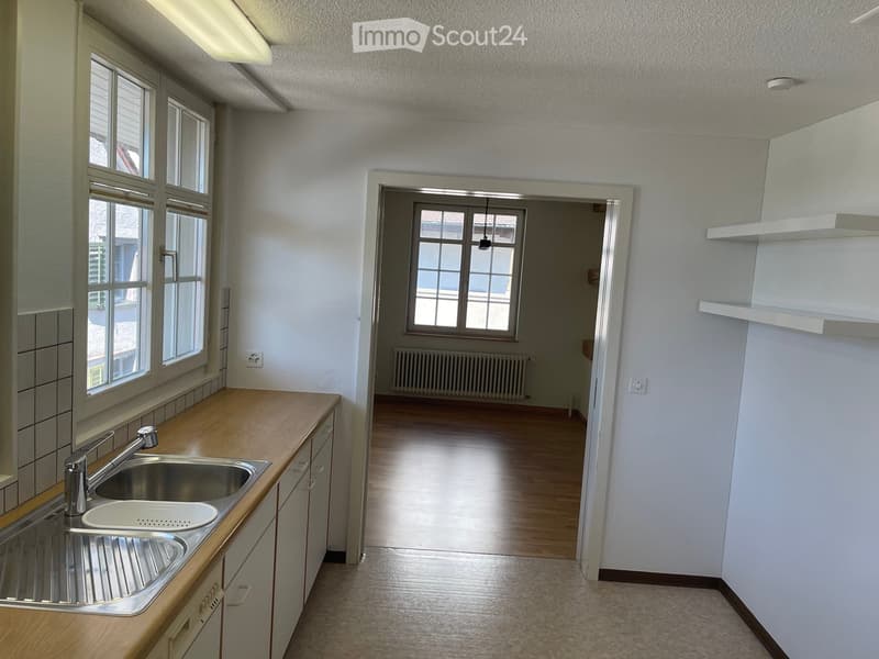 Empty kitchen with wooden countertops, stainless steel sink, and white cabinets. Adjacent room visible with wooden flooring and radiator.