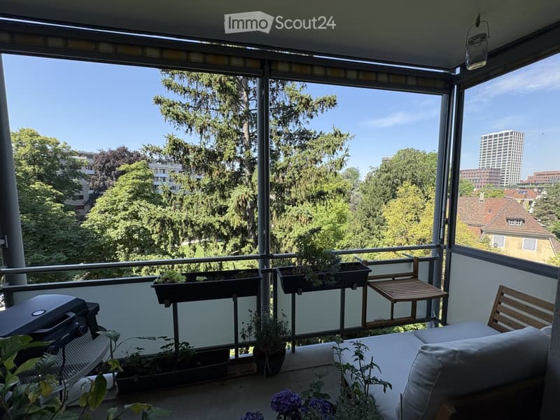 Balcony with a sofa, chair, small table, and several potted plants, offering a view of trees and a cityscape.