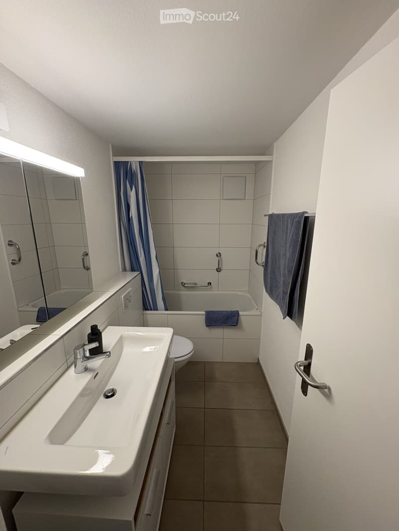 Modern bathroom with a white sink, mirror, bathtub, and toilet. Blue towels are placed on the bathtub and a shelf. The room features tiled walls and a clean, contemporary design.