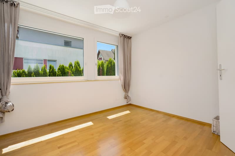 Empty room with light wooden flooring, white walls, large windows with curtains, and a small wicker basket in the corner.