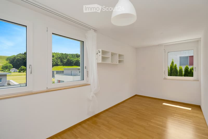 Empty room with light wooden flooring, white walls, large windows with white curtains, and a small built-in shelf unit.