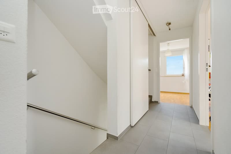 Bright, modern hallway with white walls, tiled floor, and a staircase leading upwards. A doorway leads to another room with wooden flooring and a window.