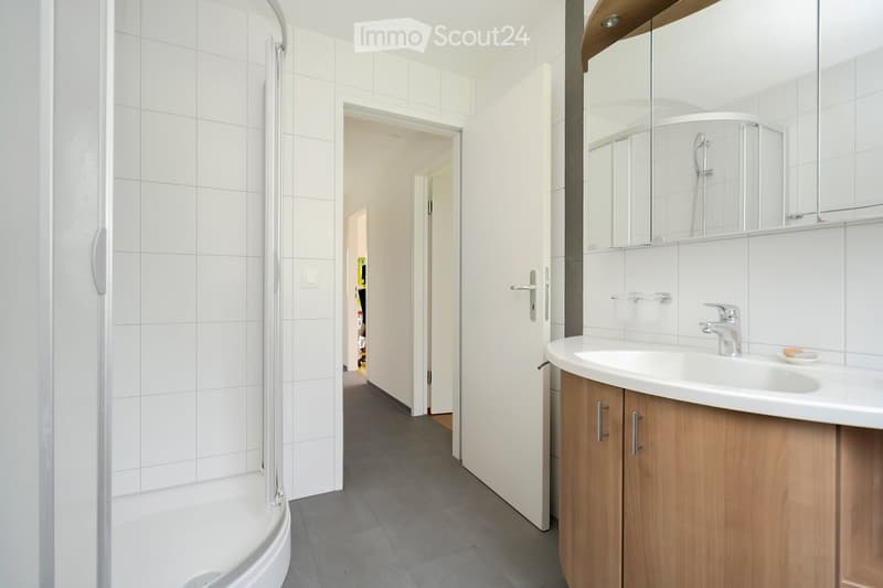 Modern bathroom with white tiled walls, a shower area, a sink with a wooden cabinet, and a large mirror above the sink.
