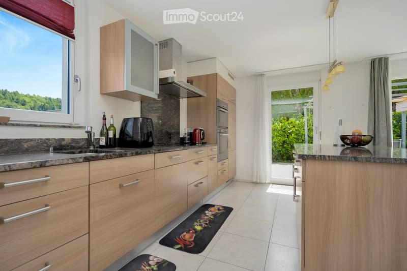 Modern kitchen with light wooden cabinets, stainless steel appliances, granite countertops, and a breakfast bar with a bowl of fruit. The space features a window with a view of greenery and a decorative rug on the floor.