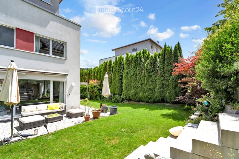 Well-maintained garden with a neatly trimmed lawn, a modern white house in the background, and outdoor seating area with a sofa, coffee table, and potted plants.