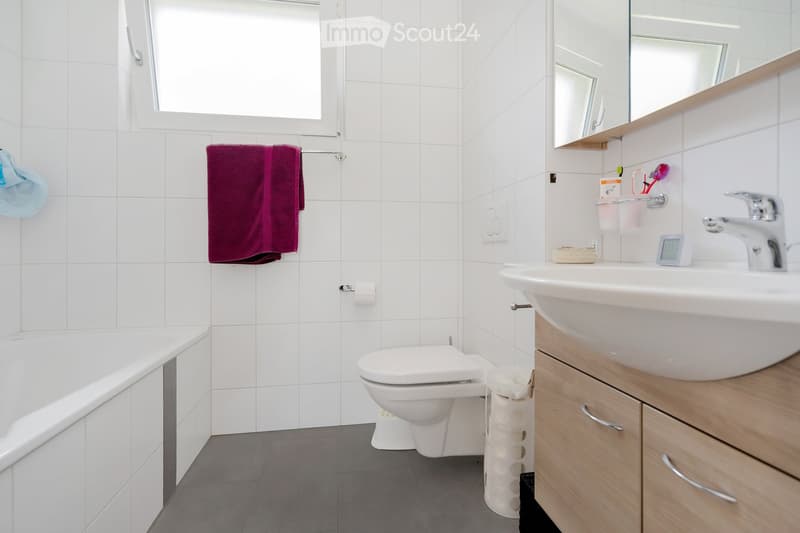 Modern bathroom with white tiles, a bathtub, toilet, sink, and a large mirror. Includes a towel rack with a red towel, a toothbrush holder, and a temperature display on the sink counter.