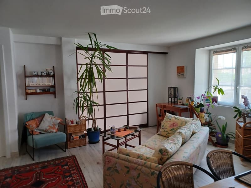 A cozy living room with a floral-patterned sofa, a teal armchair, a coffee table, a large potted plant, a shelf with books and decorative items, and a small desk with a laptop and plants.