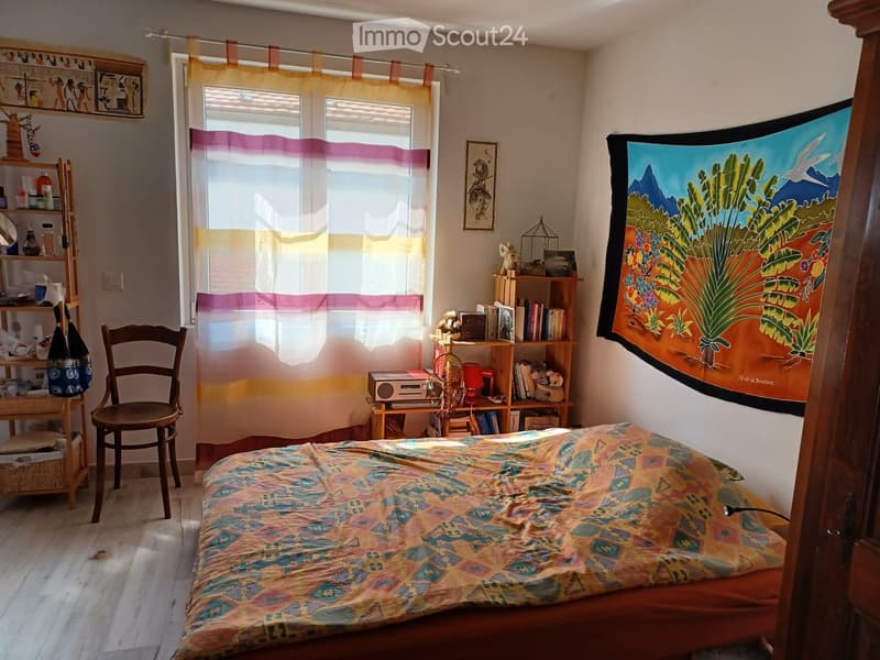 Cozy bedroom with wooden floor, large window with colorful curtains, bed with patterned bedspread, wooden chair, shelves with books and decorative items, and a colorful tapestry on the wall.