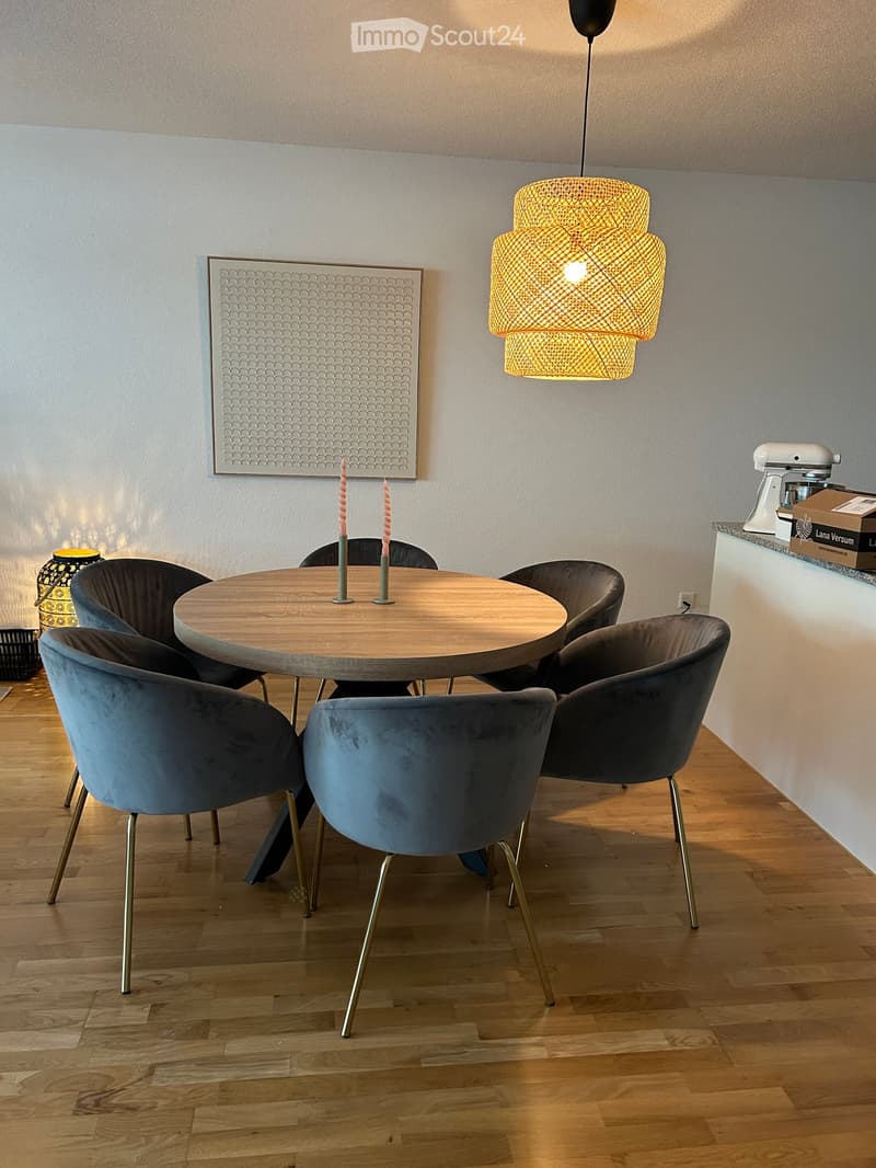 Dining area with a round wooden table surrounded by six upholstered chairs, a decorative wall art piece, and a modern hanging lamp. A small kitchen counter with a mixer is visible in the background.