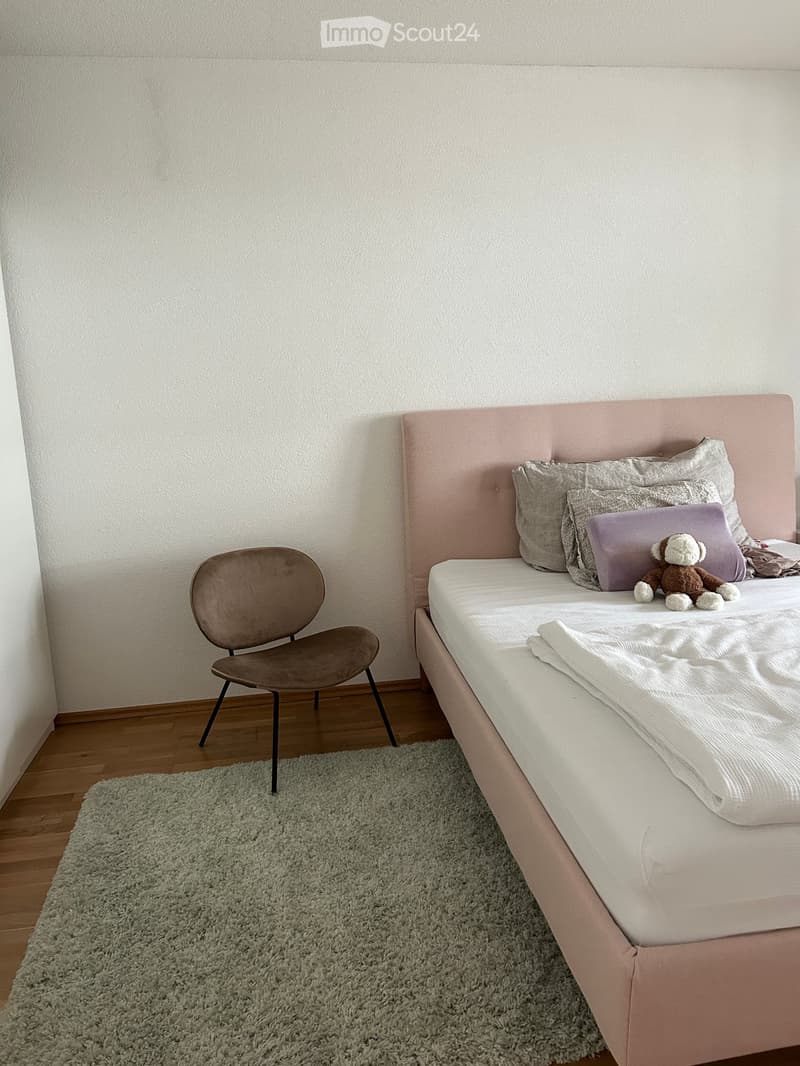 A modern bedroom with a light pink upholstered bed, grey pillows, a stuffed animal, a grey chair, and a shaggy rug on a wooden floor.