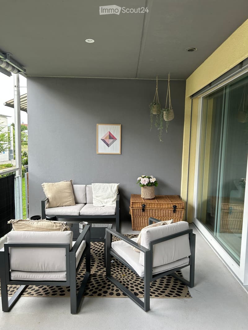Modern balcony with grey walls, white cushions, a sofa, two armchairs, a coffee table, a wicker chest, and hanging plants.