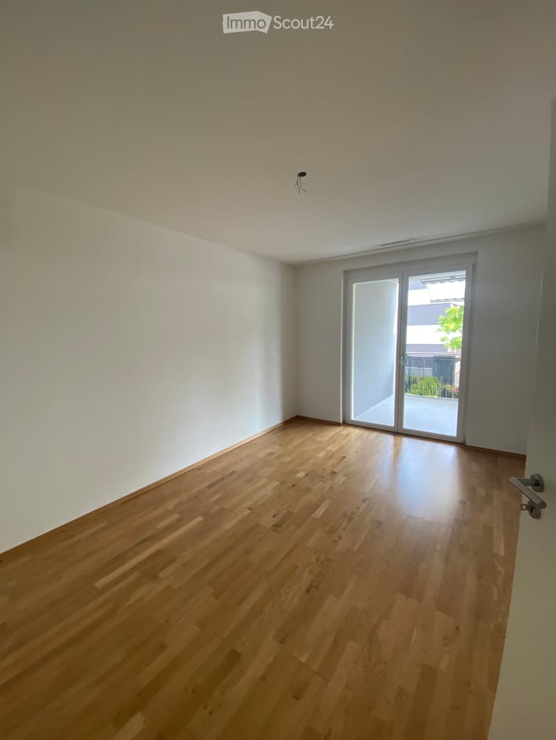Empty room with wooden flooring and white walls, featuring a sliding glass door leading to a balcony or outdoor area.