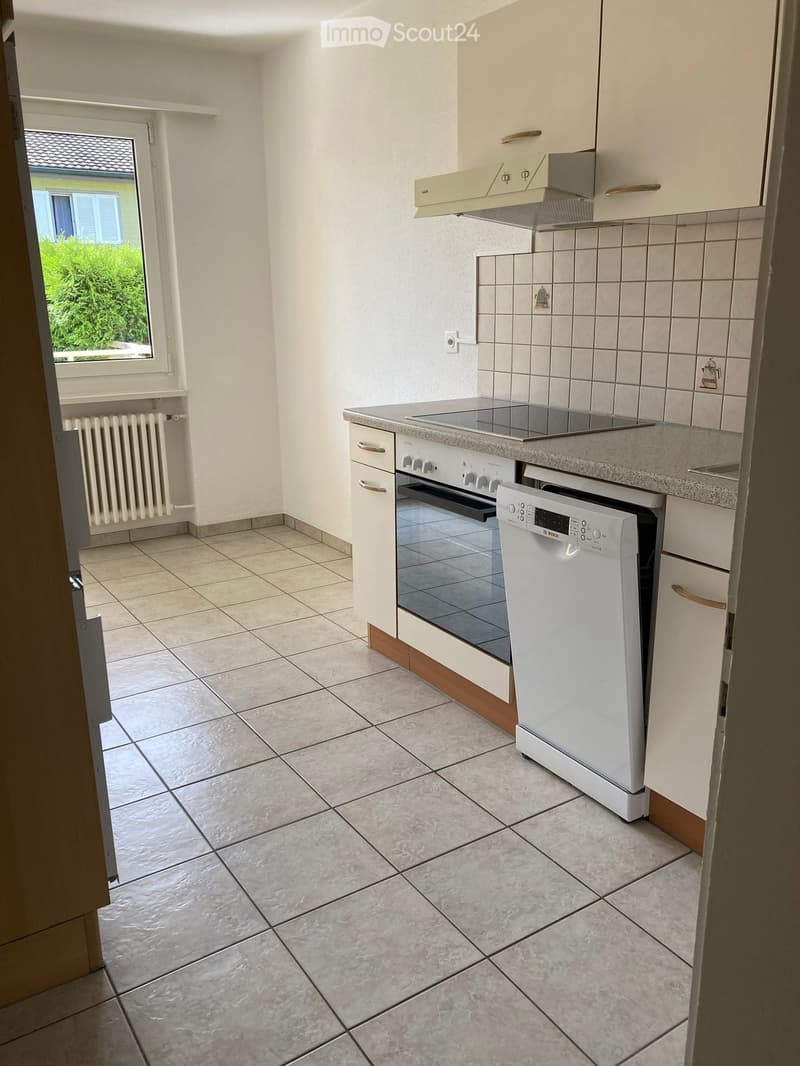 Modern kitchen with white cabinets, tiled backsplash, granite countertop, built-in oven, dishwasher, and range hood. Light-colored tiled floor and window with view of greenery.