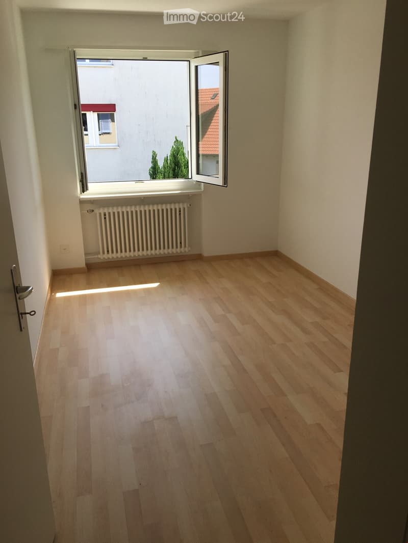 Empty room with light wooden flooring, white walls, a radiator, and an open window showing a view of neighboring buildings and trees.