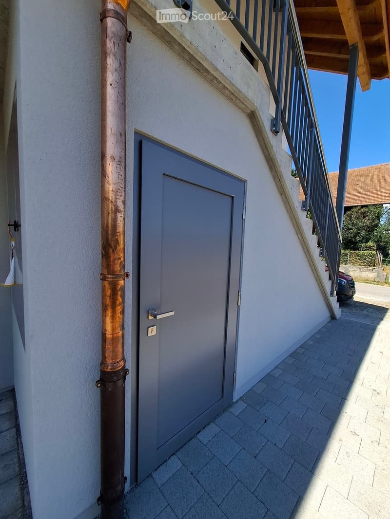 A narrow outdoor corridor with a grey door, a copper pipe, and a staircase leading upwards.