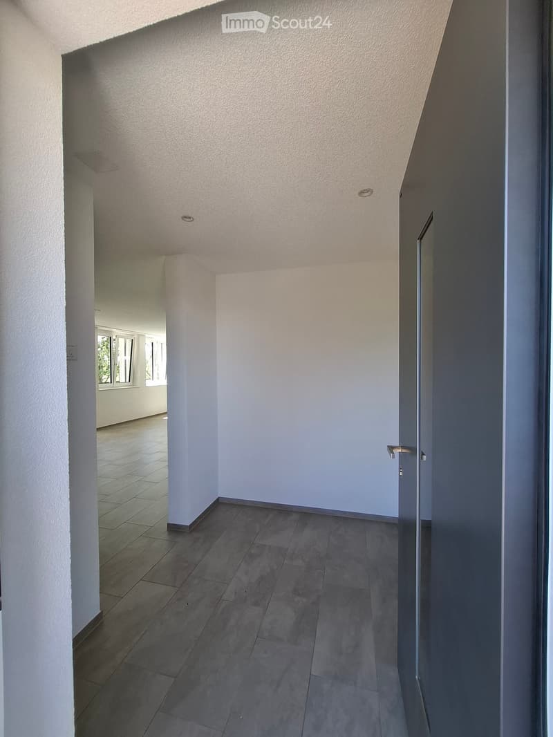 Empty hallway with tiled floor, white walls, and recessed lighting. A door is visible on the right side, and a glimpse of another room with windows is visible at the end of the hallway.