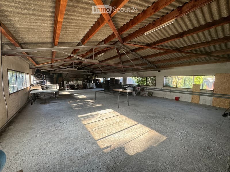 An empty, spacious room with a concrete floor and exposed wooden beams on the ceiling. The room has large windows allowing natural light to enter, and there are some storage items and equipment visible in the background.