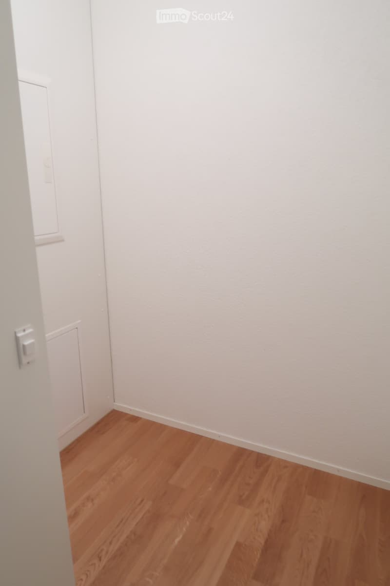 Empty room with light wooden flooring and white walls, featuring a partially visible adjacent room with a white door and light switch.
