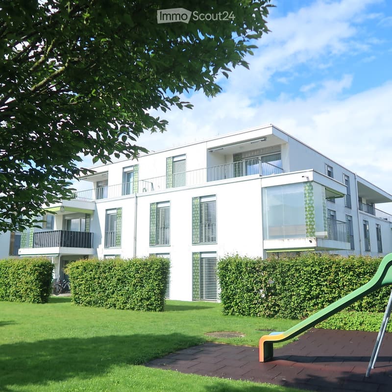 Modern white multi-story building with balconies, large windows, and greenery in the foreground.