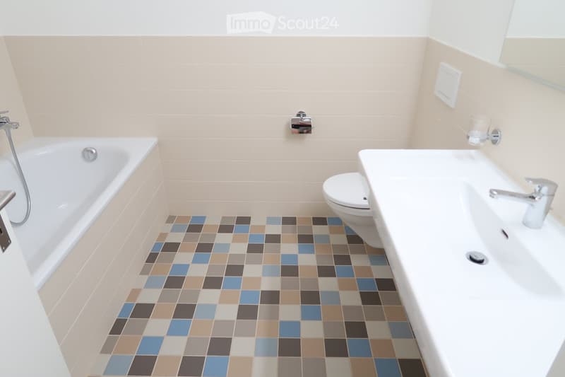 Modern bathroom with a bathtub, toilet, sink, and a colorful mosaic tile floor. The walls are tiled in a light beige color, and the fixtures are silver.
