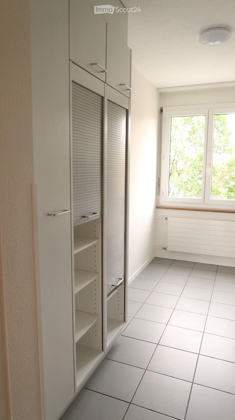 Empty room with white tiled floor, white walls, built-in shelving unit, and large windows with blinds.