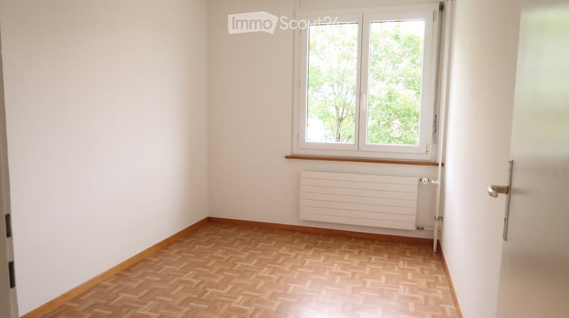 Empty room with wooden herringbone flooring, white walls, a window with white frame, and a white radiator below the window.