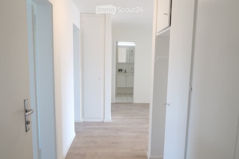 A narrow, well-lit hallway with light wooden flooring and white walls. A small, modern bathroom is visible at the end of the corridor, featuring white tiles, a sink, and a mirror.