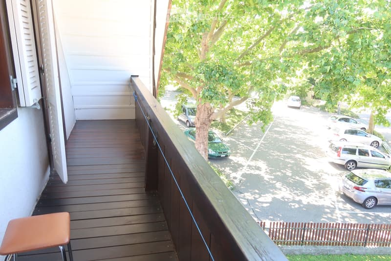 Wooden balcony, metal railing, parking area with multiple cars, grassy area with a fence, building with white walls and shutters, tree in the parking area
