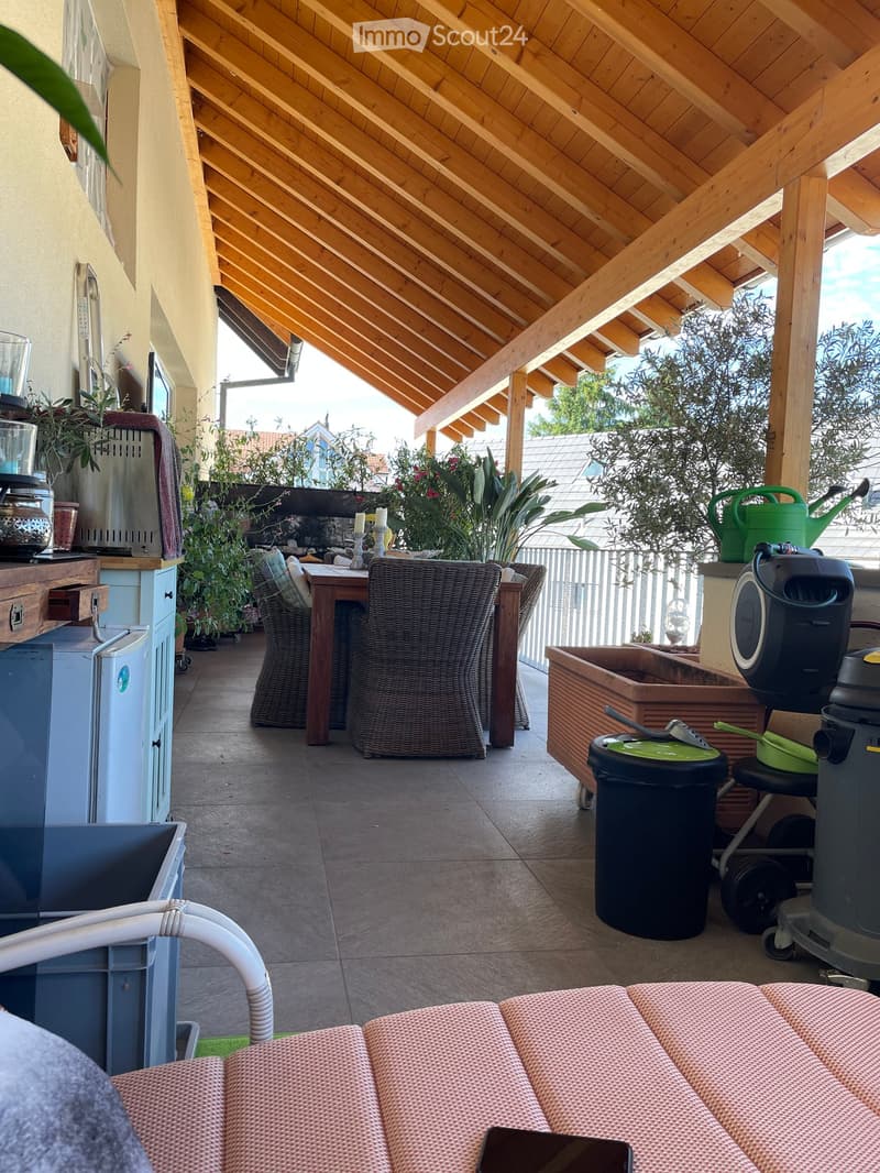 A covered outdoor patio with wooden beams, tiled floor, and outdoor furniture including a table, chairs, and a small refrigerator.